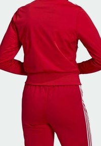 Czerwona sportowa kurtka wykonana z gładkiego materiału, o dopasowanym kroju, z prążkowanym dołem i rękawami. Posiada trzy białe paski po bokach.