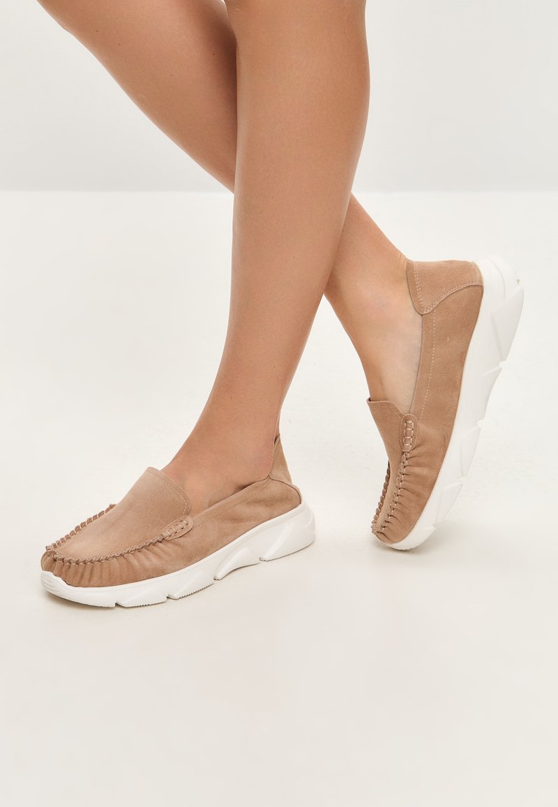 Beige suède instappers met een getextureerde franjestructuur en witte zolen, met een ronde neus en een minimalistisch ontwerp.