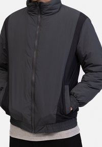 Gray zip-up jacket with a high collar, featuring contrast black side panels, elastic cuffs, and two side pockets. Smooth, lightweight fabric.