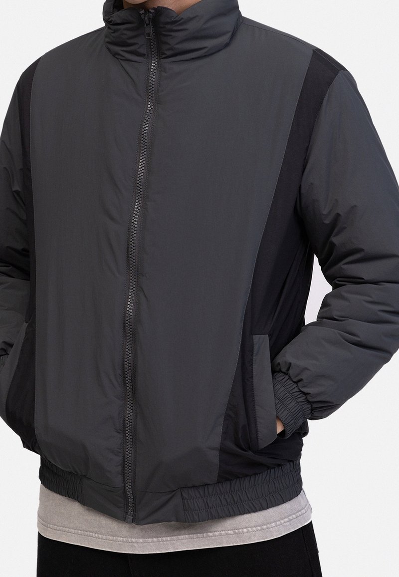 Gray zip-up jacket with a high collar, featuring contrast black side panels, elastic cuffs, and two side pockets. Smooth, lightweight fabric.