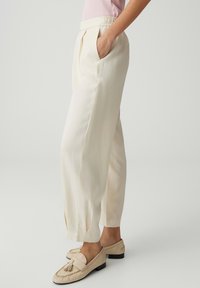 Person trägt cremefarbene, maßgeschneiderte Hosen mit elastischem Bund und beige Quasten-Slipper, steht seitlich mit einer Hand in der Tasche.