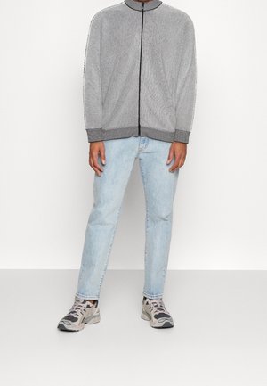 Man draagt een grijs vest met rits, lichtblauwe spijkerbroek en grijze sportschoenen, staand tegen een effen witte achtergrond.