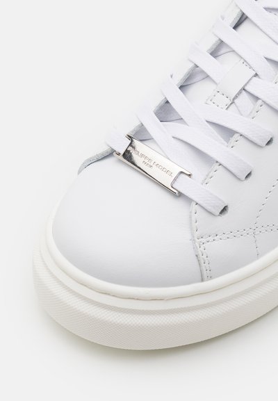 Zapatilla de cuero blanco con textura suave, presenta cordones blancos y una etiqueta de metal en tono plata con "Philippe Model" grabado. Suela de goma.
