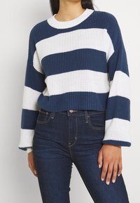 Pull tricoté avec des rayures horizontales bleues et blanches, texture côtelée, manches larges et encolure ronde, porté avec un jean foncé.