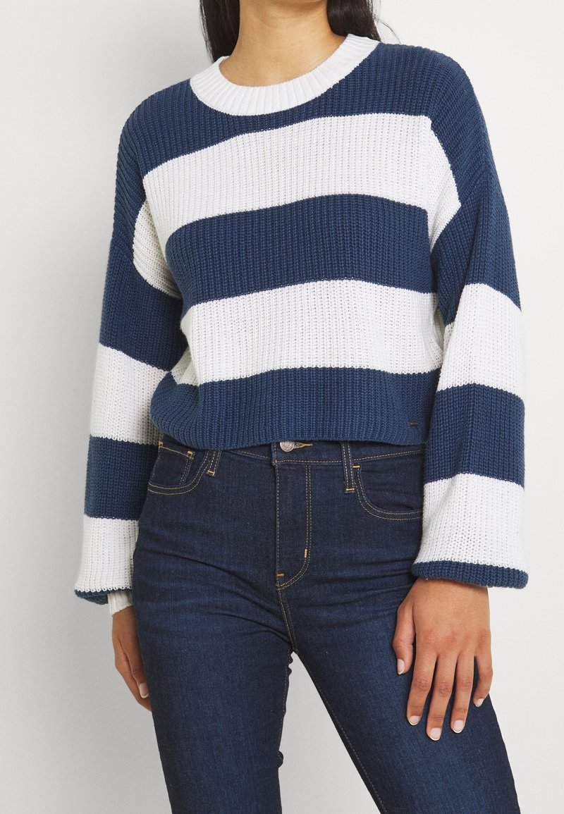 Pull tricoté avec des rayures horizontales bleues et blanches, texture côtelée, manches larges et encolure ronde, porté avec un jean foncé.