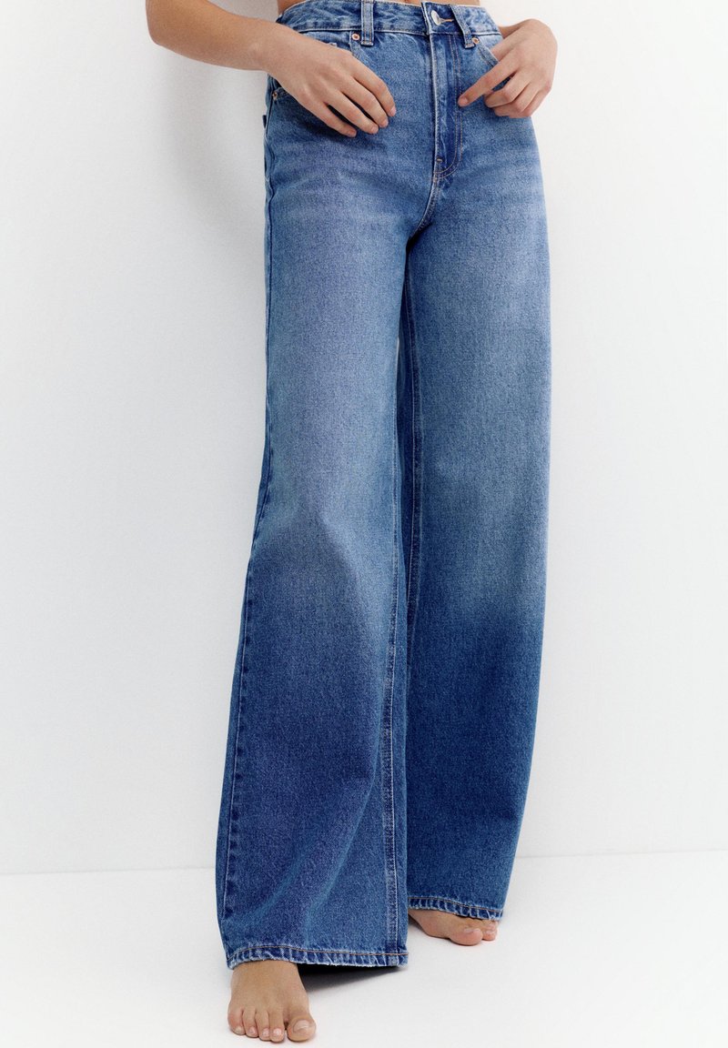 Femme portant un jean denim bleu taille haute à jambes larges, debout pieds nus contre un mur blanc.