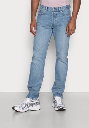 Uomo con jeans azzurri a gamba dritta e scarpe da corsa bianche con dettagli argentati, in piedi davanti a uno sfondo semplice.