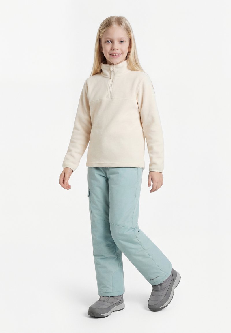 Junges Mädchen steht, trägt ein cremefarbenes Fleece-Zip-Up-Top, hellblaue Schneehose und graue Winterstiefel, lächelt in die Kamera.