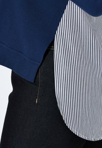 Blå sweatshirt med rundad nederkant, lager på lager över en vit och svart randig skjorta, tillsammans med mörka denimjeans. Subtil sömdetalj synlig.