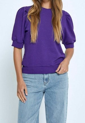 Femme portant un haut violet à manches bouffantes et un jean bleu clair, debout, une main reposant le long du corps et l'autre sur la taille.