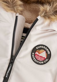 Light pink jacket with a faux fur hood, zippered front, and a circular "Superdry Expedition Co." patch featuring mountain graphics.