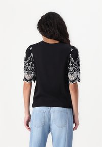 Svart kortärmad t-shirt med intrikat vitt broderi på ärmarna och rund hals, parat med ljusblå denimjeans.