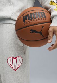 Pantalones de chándal grises con un gran logo de corazón rojo, que presenta "PUMA". Un balón de baloncesto marrón se sostiene con la mano derecha, mostrando el texto "PUMA BASKETBALL".