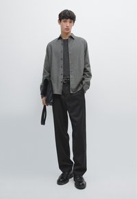 Camicia grigia a maniche lunghe sopra una t-shirt nera, abbinata a pantaloni neri a pieghe e scarpe nere. Tiene una borsa nera. Design semplice e moderno.