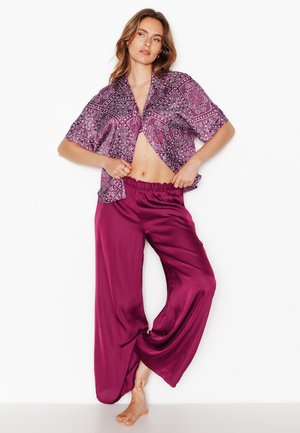 Vrouw draagt een paars, met patroon, korte mouw shirt en losse satijnen magentakleurige broek, staand op blote voeten tegen een witte achtergrond.