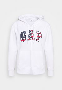 Sweat à capuche blanc zippé avec le logo "GAP" en rouge, bleu et motifs. Il dispose d'une capuche, de poches avant, et de poignets et ourlet côtelés.