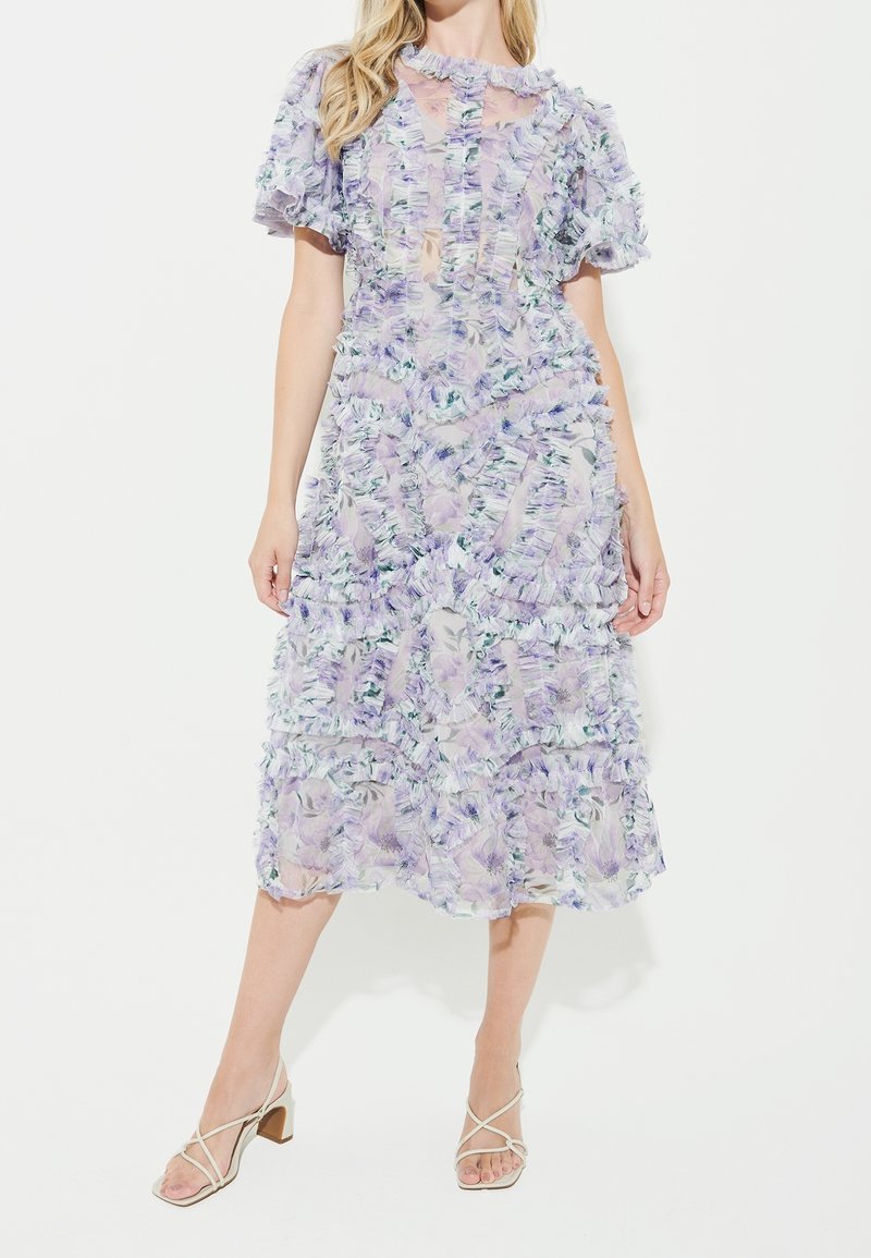 Femme portant une robe fleurie lavande transparente mi-genou avec volants et manches courtes, associée à des sandales blanches à talons blocs avec brides.