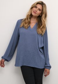 Blå, langærmet bluse med V-hals og samlet manchetter. Stoffet har en subtil tekstur og afslappet pasform, kombineret med sorte bukser.