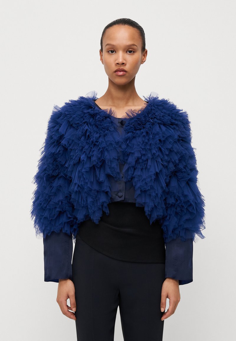 Granatowa, krótka kurtka z marszczonego tiulu, okrągły dekolt i zapięcie na guziki; w połączeniu z dopasowanym czarnym topem i ciemnymi spodniami.