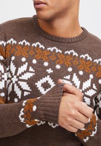Suéter de punto marrón con patrones de copos de nieve en blanco y naranja. Los puños y el cuello acanalados añaden textura y detalle al diseño.