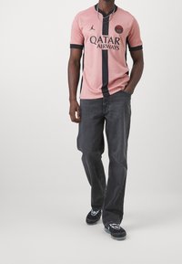 Muž v růžovém a černém dresu Fotbalového klubu Paris Saint-Germain, tmavých šedých džínách a černobílých teniskách, jde s rukou v kapse.