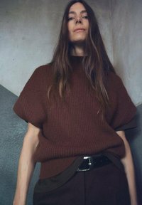 Suéter marrón de punto con mangas cortas y un cuello alto, combinado con pantalones oscuros y un cinturón negro, frente a un fondo gris texturizado.