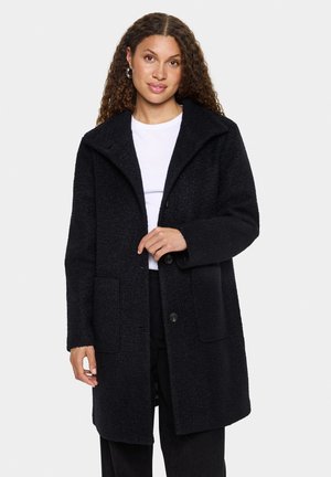 Black textured coat with a wide collar, front button closure, and side pockets. Worn over a white top, features long sleeves and a relaxed fit.