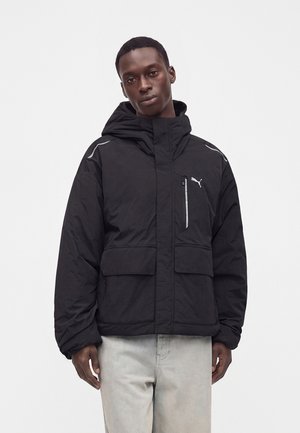 Jeune homme portant une veste à capuche noire Puma avec de grandes poches avant et un pantalon gris clair sur un fond blanc uni.