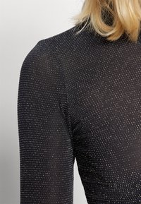 Mustapohjainen, ribbed pitkähihainen paita korkealla kaula-aukolla, jossa on hienovaraisia metallipilkkuja tekstuurin luomiseksi. Sujuva pinta pehmeässä kankaassa.