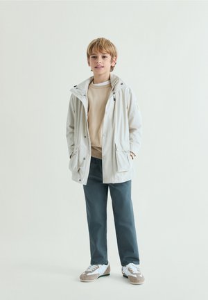Garçon debout portant une veste beige clair, un pull marron clair, un pantalon bleu foncé et des baskets blanches sur un fond clair uni.