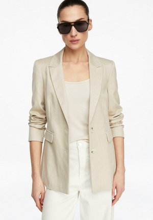 Kobieta ubrana w beżową marynarkę na beżowym topie, białe spodnie i duże czarne okulary przeciwsłoneczne, stojąca na białym tle.