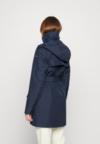 Veste imperméable bleu marine avec col montant, taille ceinturée et capuche. Caractérisée par un tissu lisse et brillant ainsi qu'un détail logo discret sur l'épaule.
