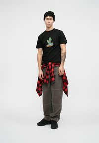 Melna T-kreklu ar zaļu grafiku, pelēkas bikses un melnas kedas. Sarkana un melna rūtainā krekla apkārt jostasvietai. Melns beanie cepure.