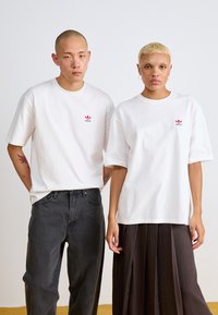 Balti medvilniniai marškinėliai su raudona ir žalia Adidas logotipu, derinami su tamsiais džinsais ir raukšlėta ruda sijonu, turintys trumpas rankoves ir laisvą siluetą.