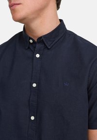 Marineblå kortermet skjorte med krage, hvite knapper og et lite brodert design på brystet. Laget av bomull, med glatt tekstur.
