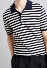Muž v modro-bílem pruhovaném polo tričku s krátkým rukávem a žebrovaným vzorem, v černých kalhotách, s rukama v kapsách.