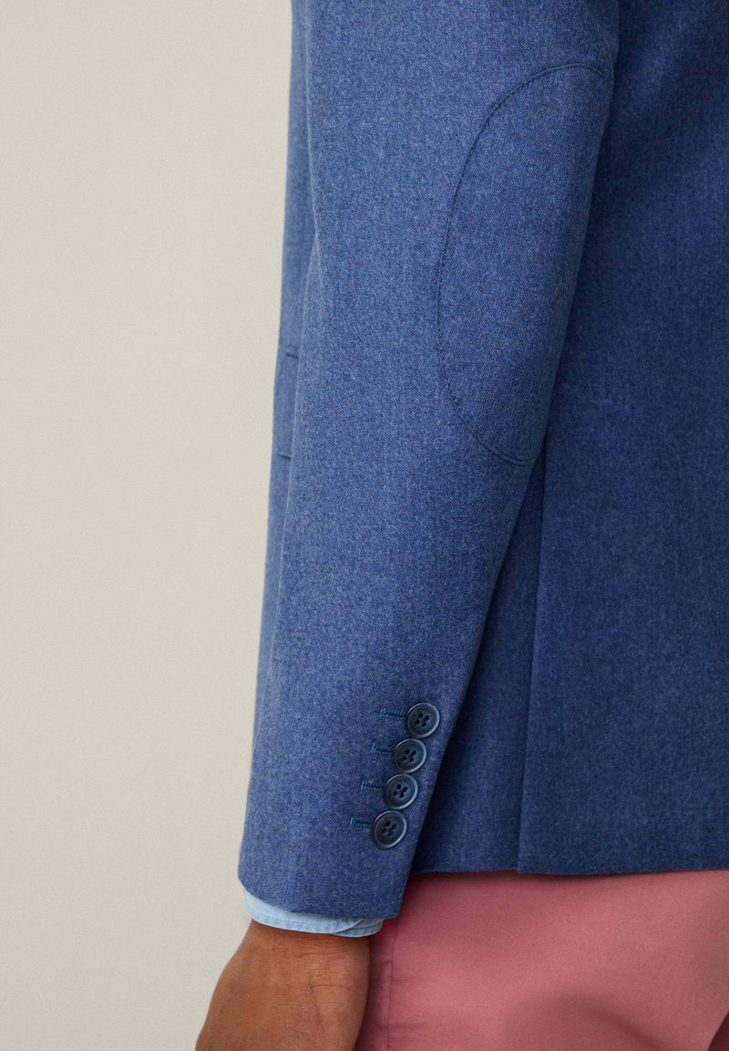 Blauwe blazer mouw met vier knopen en een elleboogpatch over een lichtblauw shirt, gedragen met roze broeken op een donkerhuidige arm.