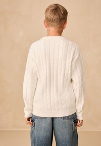 Maglione bianco lavorato a trecce con scollo tondo e orlo a coste. Presenta maniche lunghe e un motivo a rilievo sulla schiena.