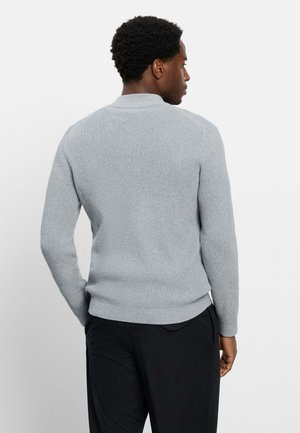 Mann mit kurzen lockigen Haaren, der einen hellgrauen strukturierten Langarmpullover und schwarze Hosen trägt, mit dem Rücken zu einem schlichten weißen Hintergrund.