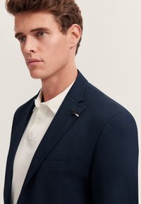 Blazer navy realizzato in tessuto liscio, con una tasca sul petto e un'icona a spillo. Indossato sopra una polo di colore chiaro.