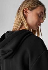 Felpa nera con cappuccio caratterizzata da una texture morbida, un ampio cappuccio e maniche raglan. Il focus è sulla linea del collo e sull'area delle spalle.