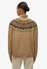 Pull beige tricoté avec un col montant, présentant un empiècement à motifs marron et blanc. Tissu texturé avec des mouchetures. Coupe ample.