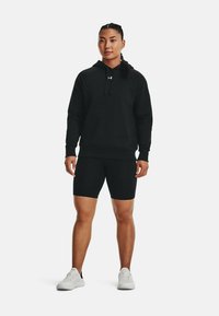 Hoodie noir avec poche devant, associé à un short noir. Porté avec des chaussures de sport blanches. Texture lisse, design simple et sans motifs.