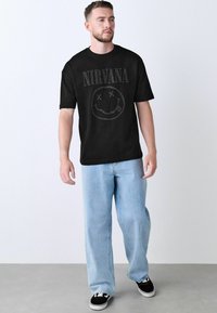 Black cotton t-shirt featuring "NIRVANA" text and a smiley face graphic with X eyes. Worn with light blue denim trousers and black sneakers.