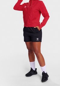 Rode rits-hoodie met een kangoeroezak, gecombineerd met zwarte shorts met een wit logo. Witte sportsokken en zwarte sneakers maken de look compleet.