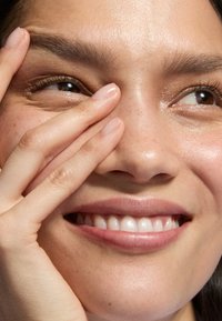 Nahaufnahme eines lächelnden Gesichts mit glatter, strahlender Haut; Hand, die teilweise das Auge bedeckt; natürliche Nägel; leichte Sommersprossen.