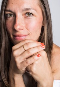 Egy női arc közeli képe, hosszú barna hajjal és mogyoróbarna szemekkel. Összeilleszti a kezeit, narancssárga körömlakkal és egy aranygyűrűvel.