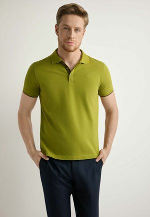 Mężczyzna z jasnobrązowymi włosami stoi z rękami w kieszeniach, ubrany w zieloną koszulkę polo i ciemne spodnie na jednolitym tle.