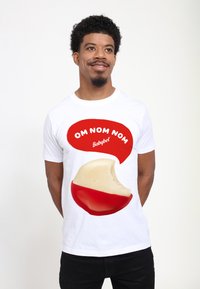 Vit t-shirt med en stor grafik av en delvis uppäten Babybel-ost med röd skorpa och texten "OM NOM NOM" i en röd pratbubbla.