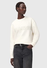 Sweter z okrągłym dekoltem w kremowym kolorze z teksturowanym logo "ALLSAINTS" ozdobionym małymi cyrkoniami. Połączony z czarnymi dżinsami o wysokim stanie.